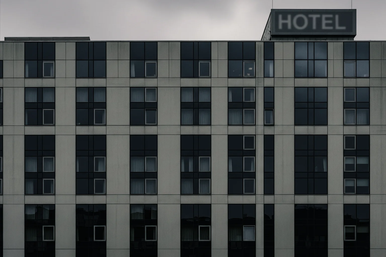 The grey, concrete facade of an anonymous modern hotel, showing a repeating grid of dark windows under an overcast sky.
