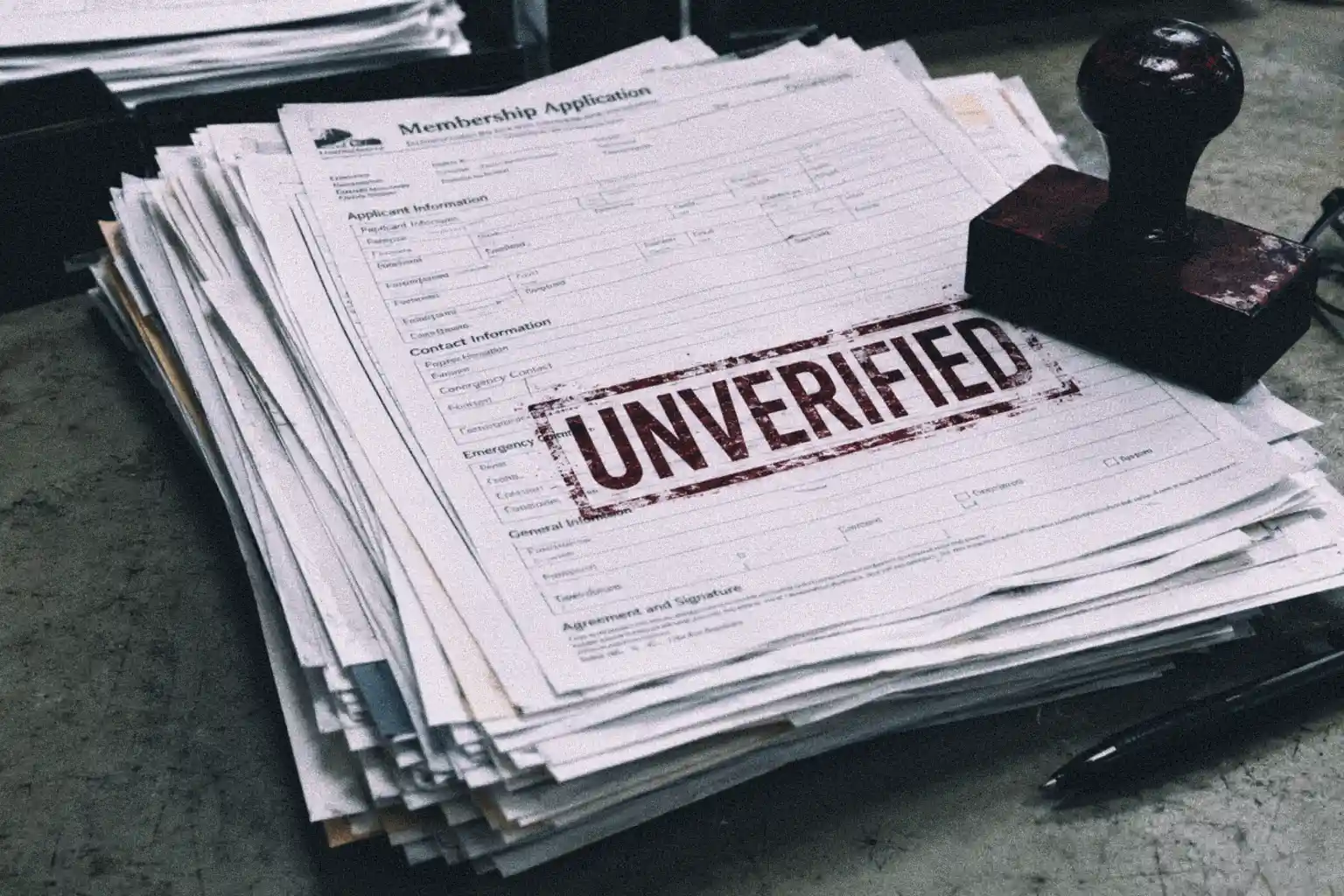 A close-up photograph of a messy stack of printed membership application forms on a desk, with the top sheet stamped 'UNVERIFIED' in red ink.
