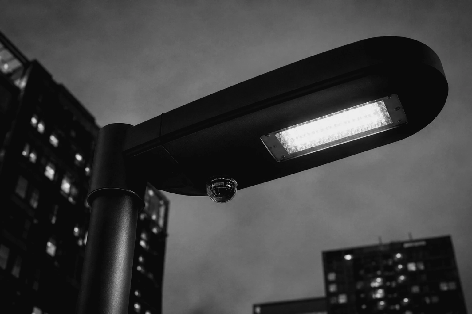 A low-angle view of a modern smart streetlight against a twilight sky. Beneath the main LED lantern sits a small glass sensor dome reflecting a grid of digital telemetry lines.